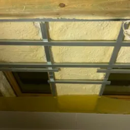 Piana izolacyjna między krokwiami dachu, widoczne metalowe profile konstrukcyjne i okna dachowe, zabezpieczone folią. Fragment instalacji elektrycznej.