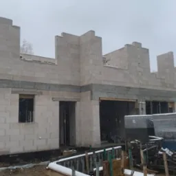 Surowy stan budynku murowanego z betonu komórkowego w zimowej scenerii, widoczne otwory okienne i drzwiowe, zbrojenie wieńca oraz materiały budowlane zabezpieczone folią