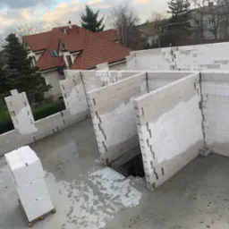 Wznoszone ściany z białych bloczków na surowym betonowym fundamencie, widoczne palety z materiałem budowlanym i fragment budynku w tle, w pochmurny dzień.