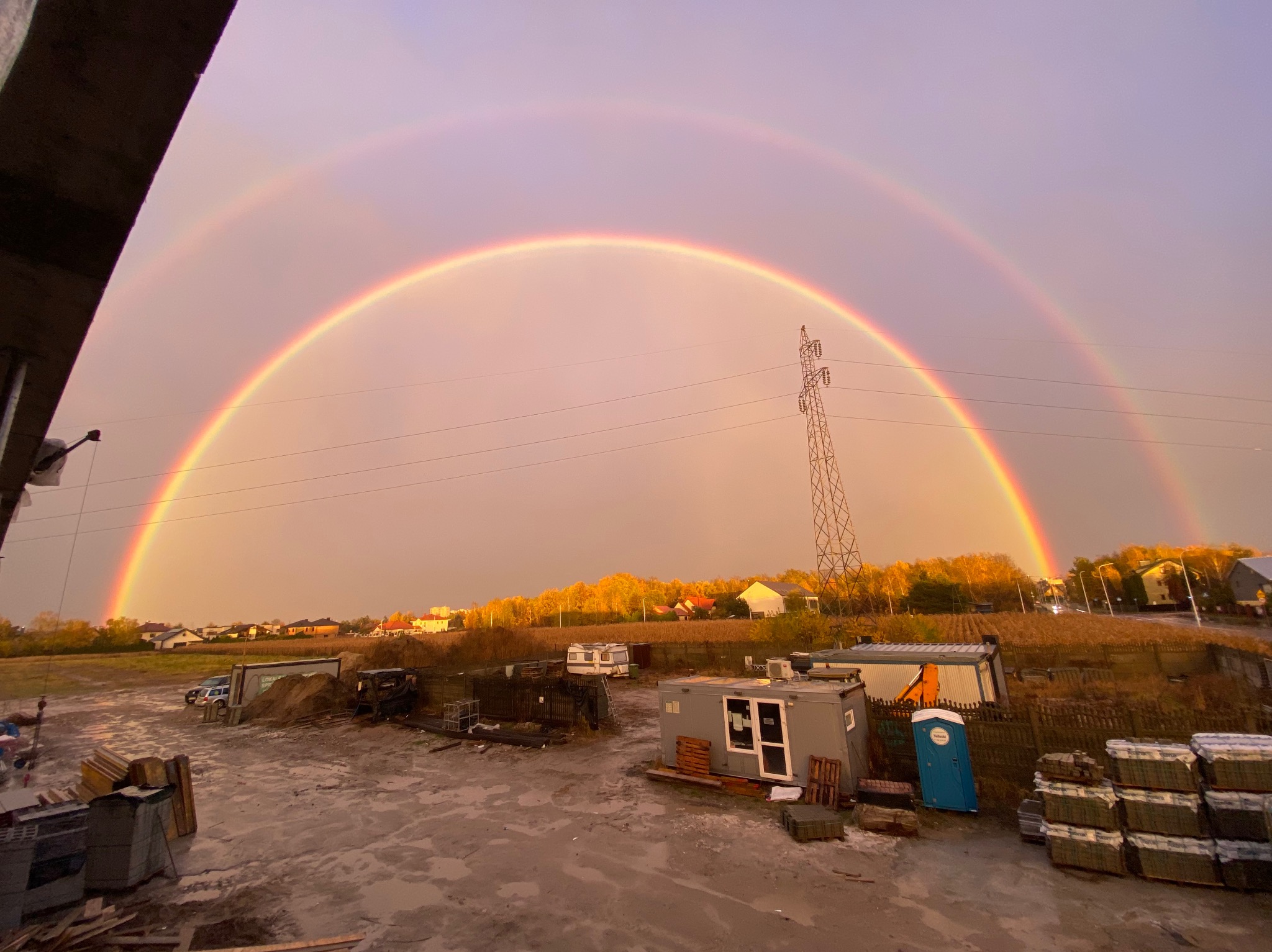 Plac budowy z barakami i stertami materiałów pod podwójną tęczą, widok z góry. Słup wysokiego napięcia w tle, jesienne drzewa i zabudowania.