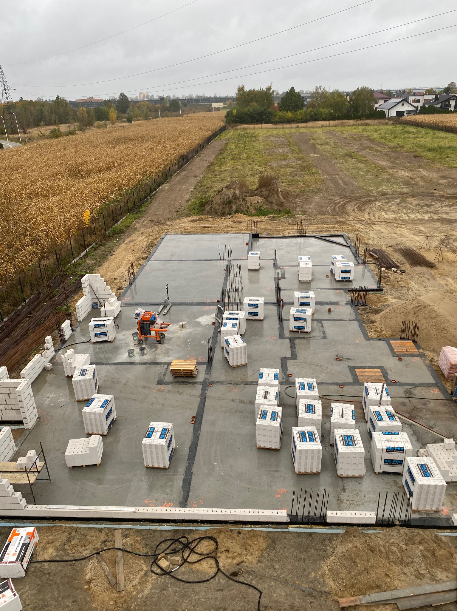 Zalana płyta fundamentowa z wystającymi prętami zbrojeniowymi i paletami bloczków SIL-PRO, widok z góry na plac budowy w pochmurny dzień.