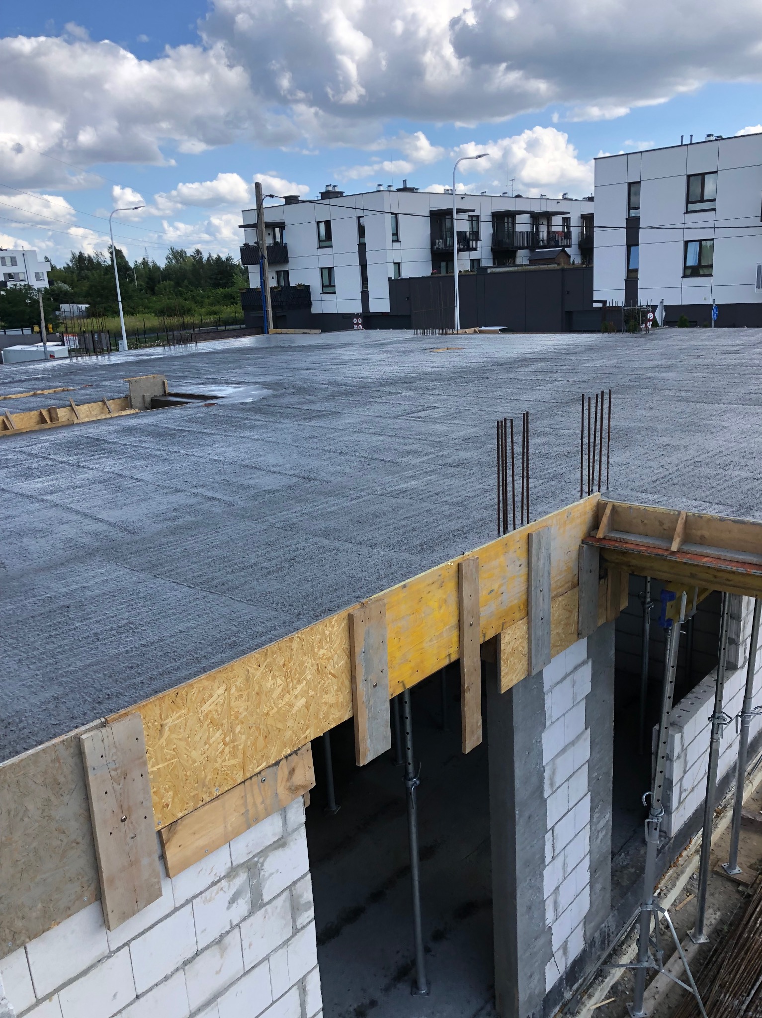 Wylana płyta fundamentowa z wystającymi prętami zbrojeniowymi, szalunkiem i fragmentem muru z białych bloczków, na tle zabudowy wielorodzinnej i błękitnego nieba.