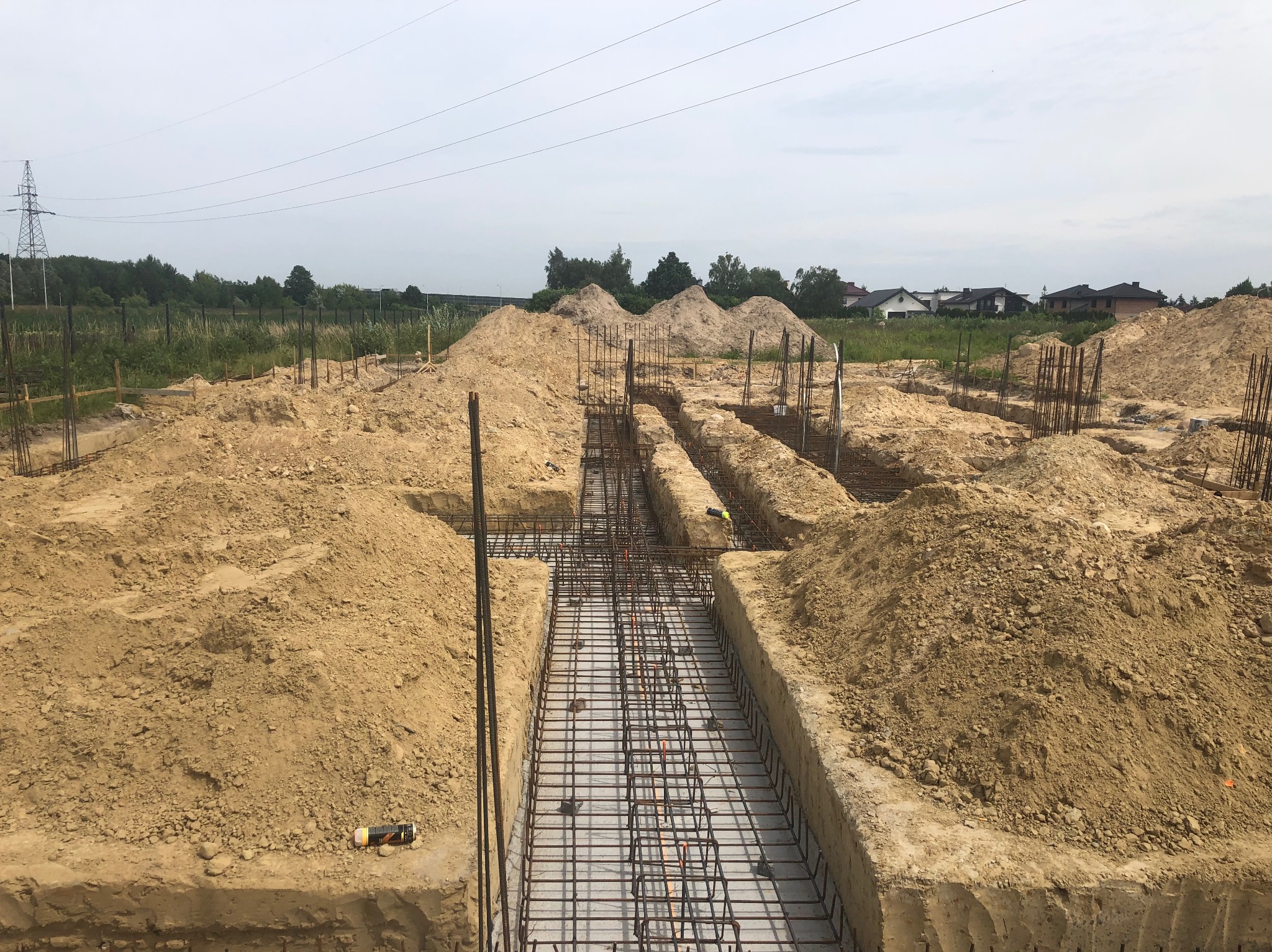 Szeroki kadr na wykopane fundamenty z ułożonym zbrojeniem, przygotowane do zalania betonem. W tle hałdy ziemi i zabudowa jednorodzinna.