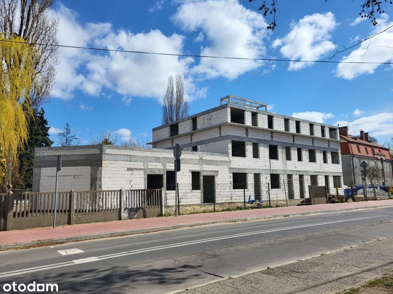 Wznoszony budynek z białych bloczków, widoczny z ulicy w słoneczny dzień. Konstrukcja w stanie surowym, z otworami okiennymi i elementami szalunku na dachu.