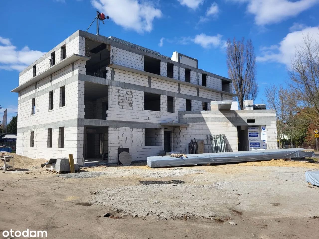 Wznoszony budynek z białych bloczków, widoczne otwory okienne i drzwiowe, na placu budowy materiały budowlane i żuraw. Ujęcie w słoneczny dzień.