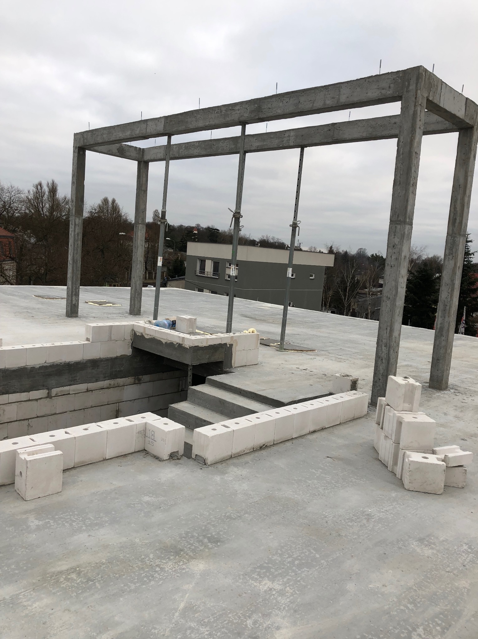 Wznoszenie konstrukcji: betonowy szkielet budynku w trakcie budowy, częściowo wymurowane ściany z białych bloczków, widok na okolicę w tle.