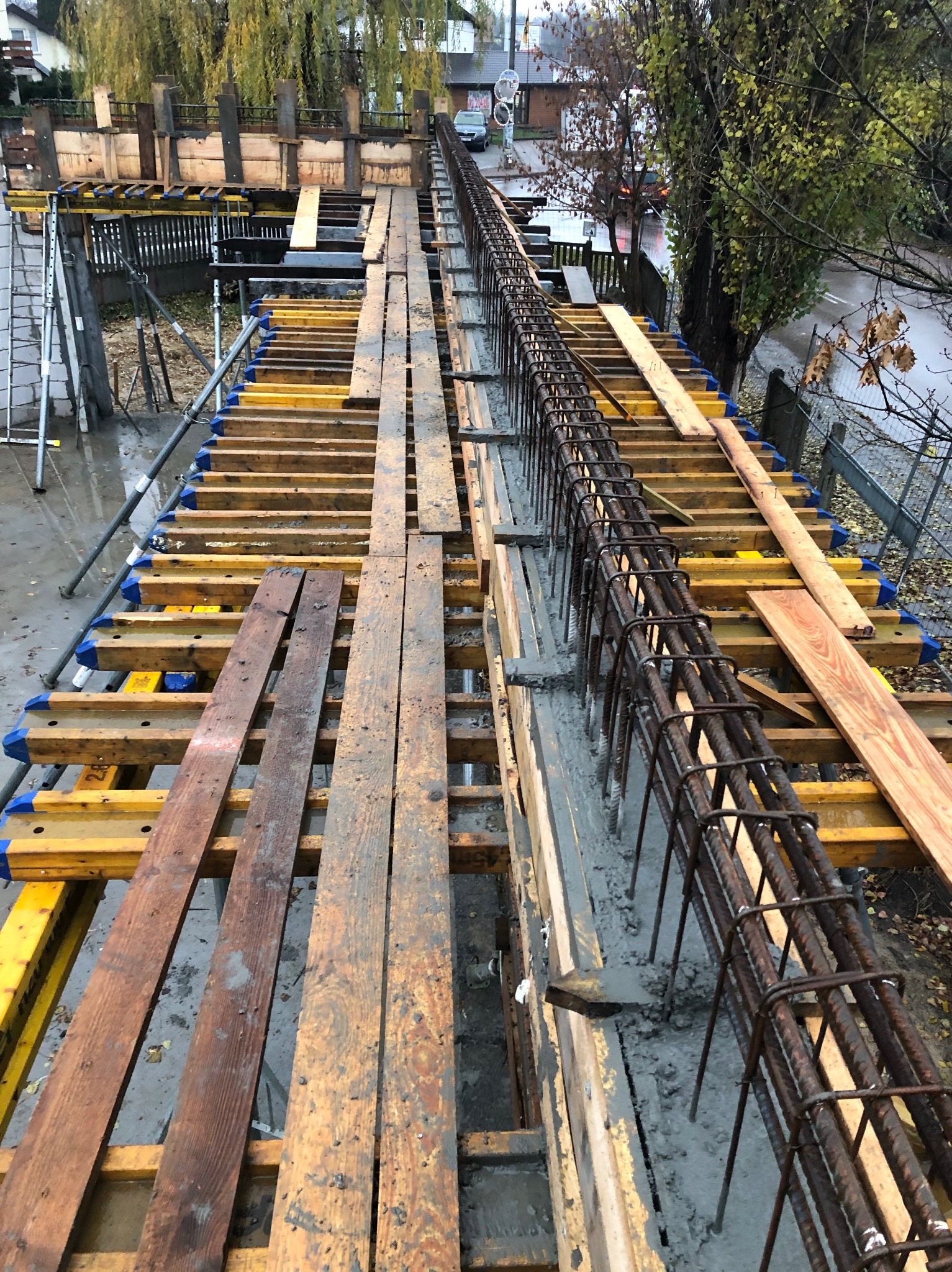 Z góry: Zbrojenie zalane betonem w szalunku z desek i żółtych podpór. Widoczne drzewa w tle. Etap budowy konstrukcji żelbetowej w pochmurny dzień.