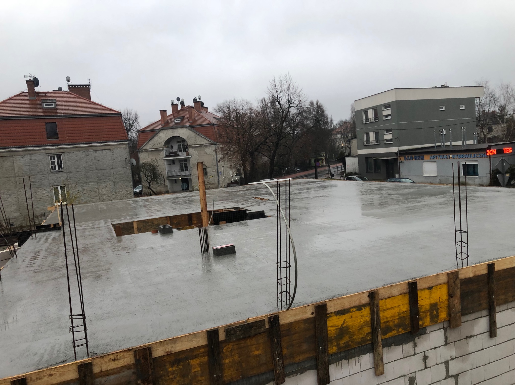 Zalana płyta fundamentowa z wystającymi prętami zbrojeniowymi, widok z góry na budowę w pochmurny dzień. Żółte szalunki i bloczki betonowe w dolnej części kadru.