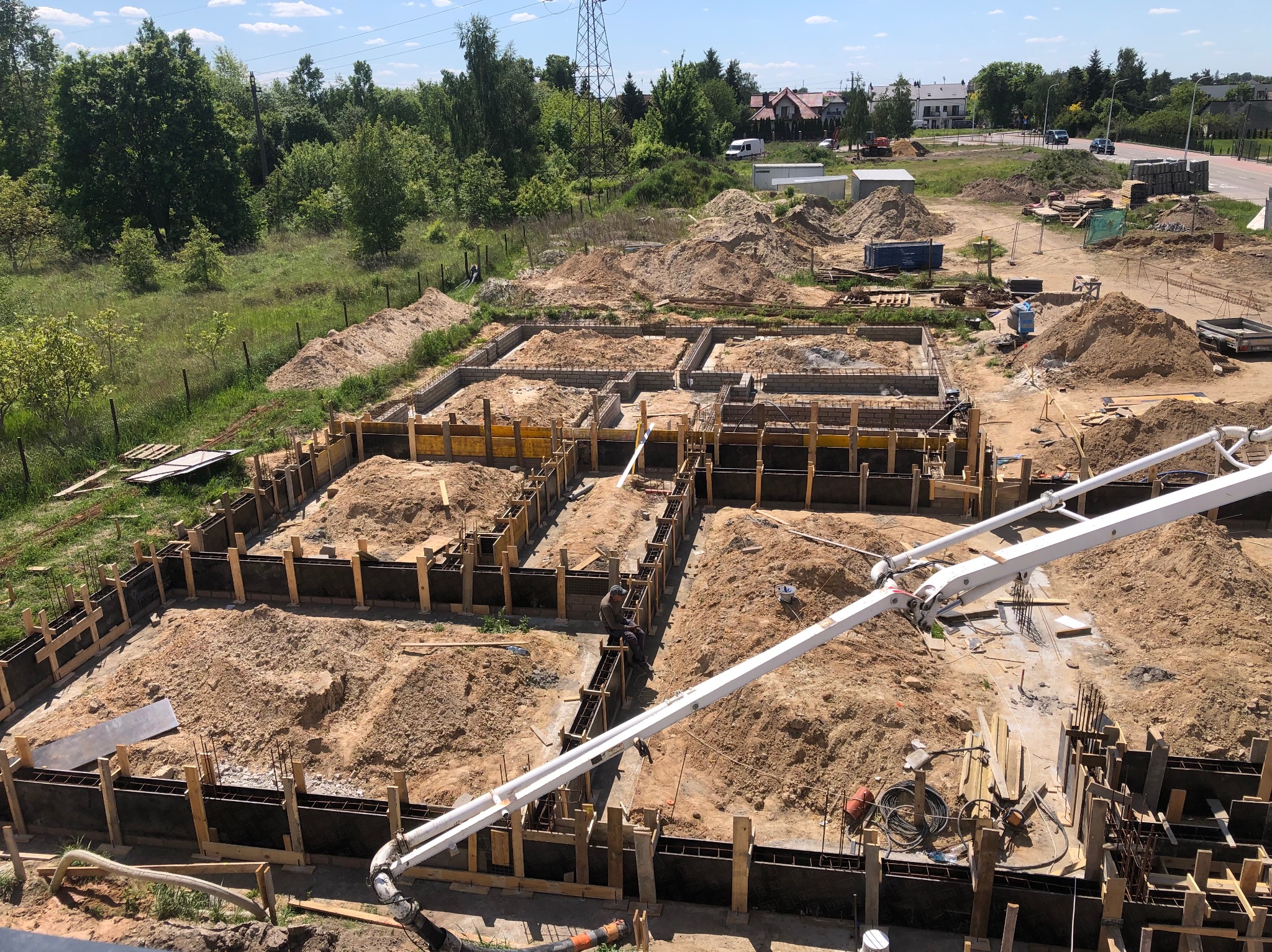 Zalewanie fundamentów domu jednorodzinnego z użyciem pompy do betonu. Widok z góry na szalunki i zbrojenie. W tle drzewa i zabudowa.