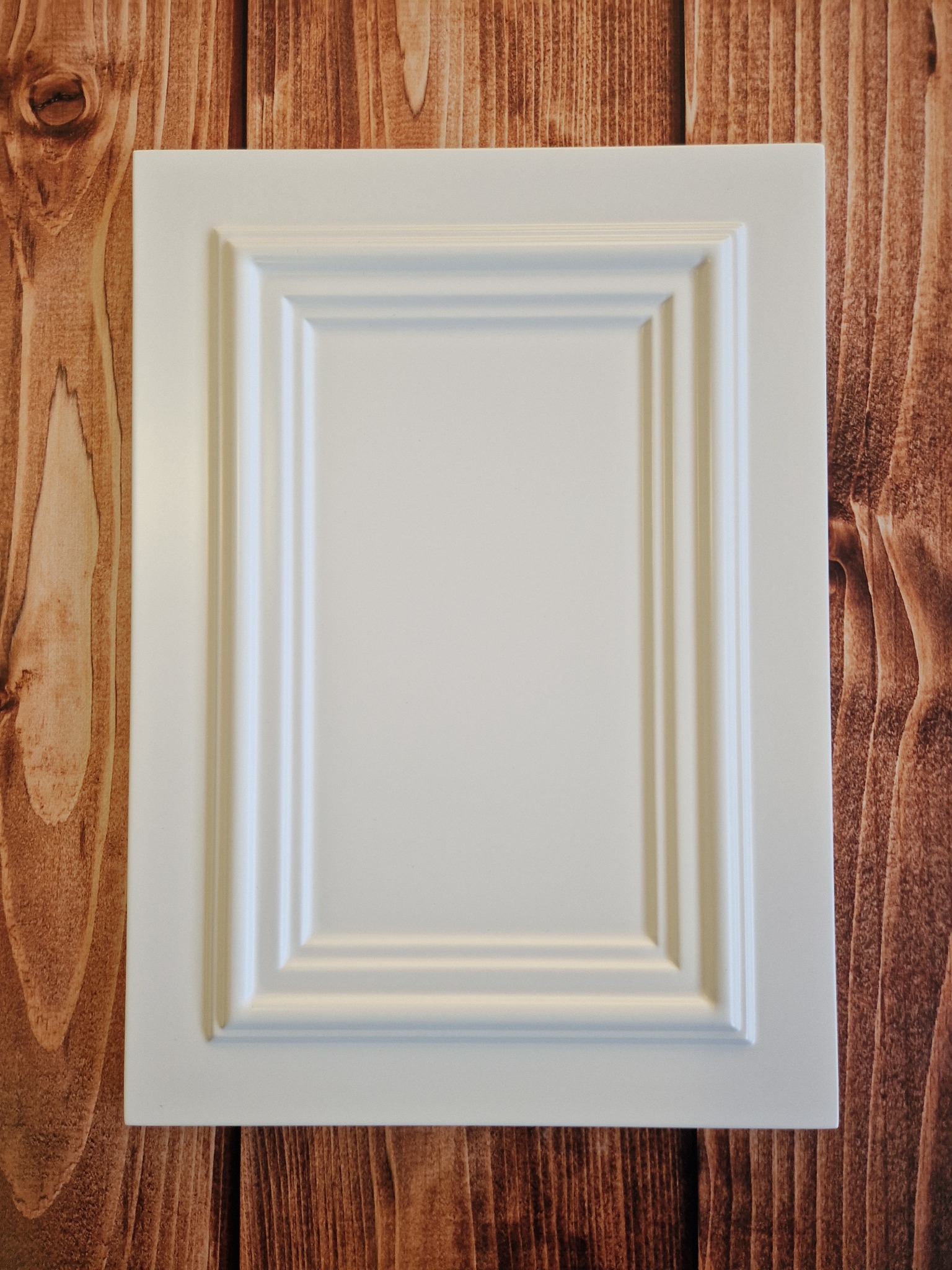 Biały, frezowany front meblowy z wyraźnym obramowaniem, leżący na tle drewnianego blatu o ciepłym, brązowym odcieniu z widocznym usłojeniem.