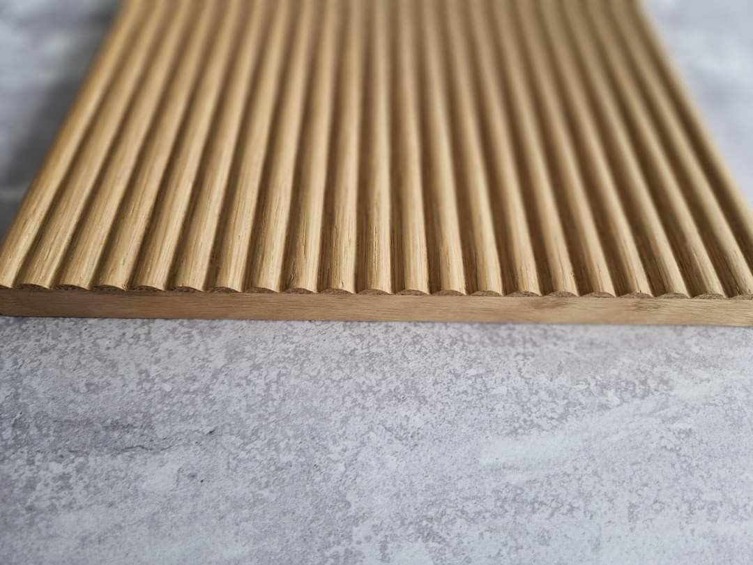 Drewniany blat z równoległymi, zaokrąglonymi żłobieniami, ukazujący naturalne usłojenie drewna na tle szarej, teksturowanej powierzchni.