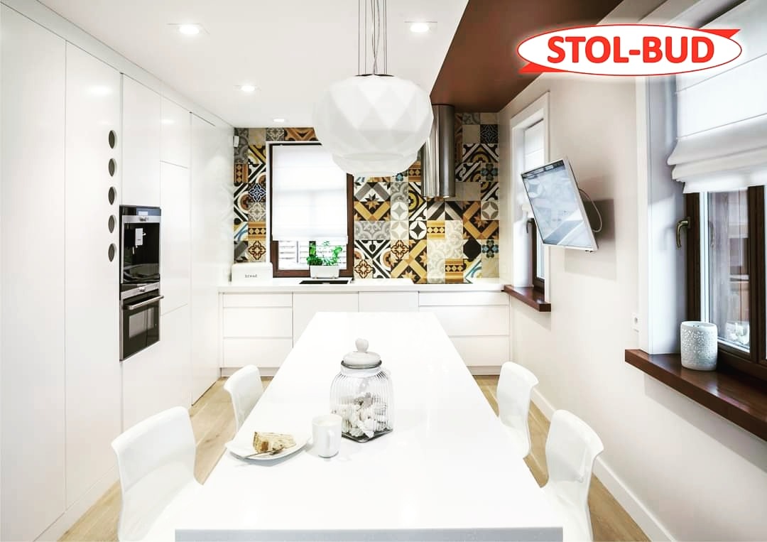 Nowoczesna, biała kuchnia na wymiar z wzorzystą ścianą nad blatem, oświetleniem punktowym i wiszącą lampą geometryczną, z widocznym logo firmy STOL-BUD.