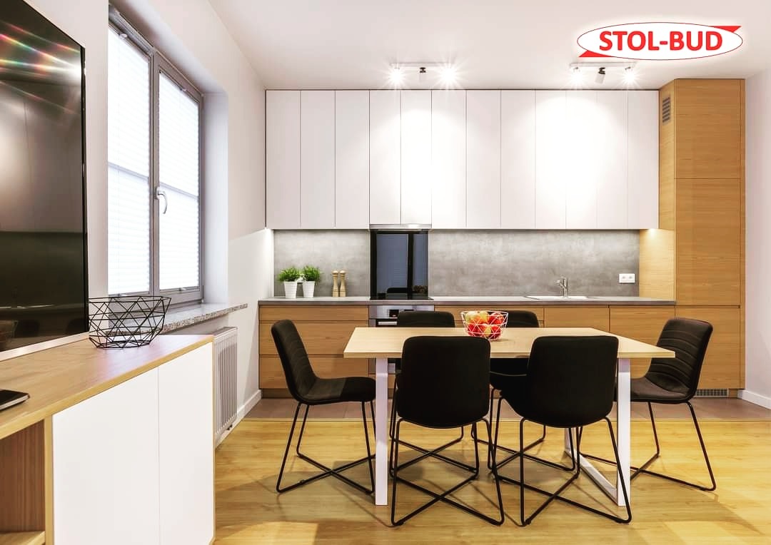 Nowoczesna kuchnia z białymi szafkami górnymi i drewnianymi dolnymi, jasny blat kuchenny, stół z czarnymi krzesłami w minimalistycznym stylu, logo firmy w prawym górnym rogu.