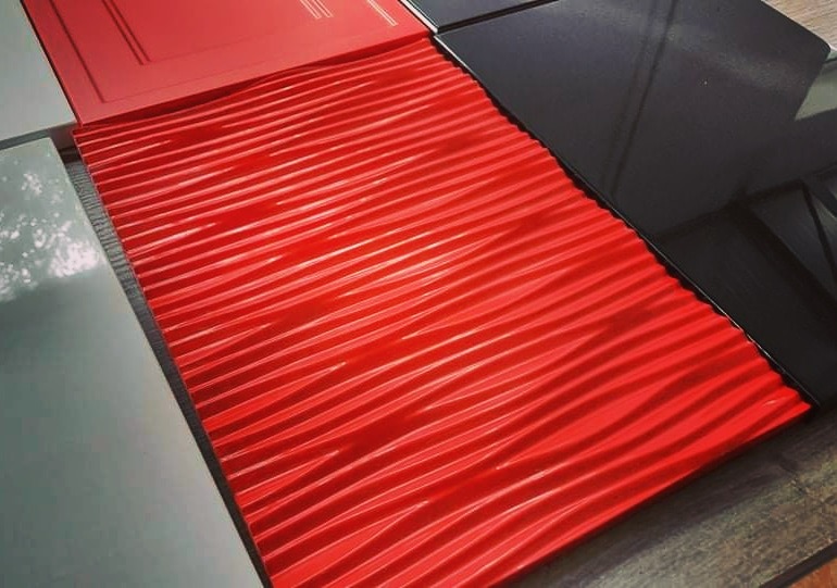 Próbki materiałów: czerwony panel z falistą fakturą, biały i czarny błyszczący blat, ułożone na drewnianej powierzchni.