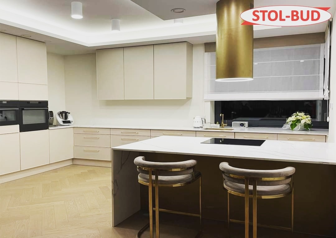 Jasna kuchnia z wyspą i hokerami na złotych nogach, z logo firmy STOL-BUD w prawym górnym rogu.