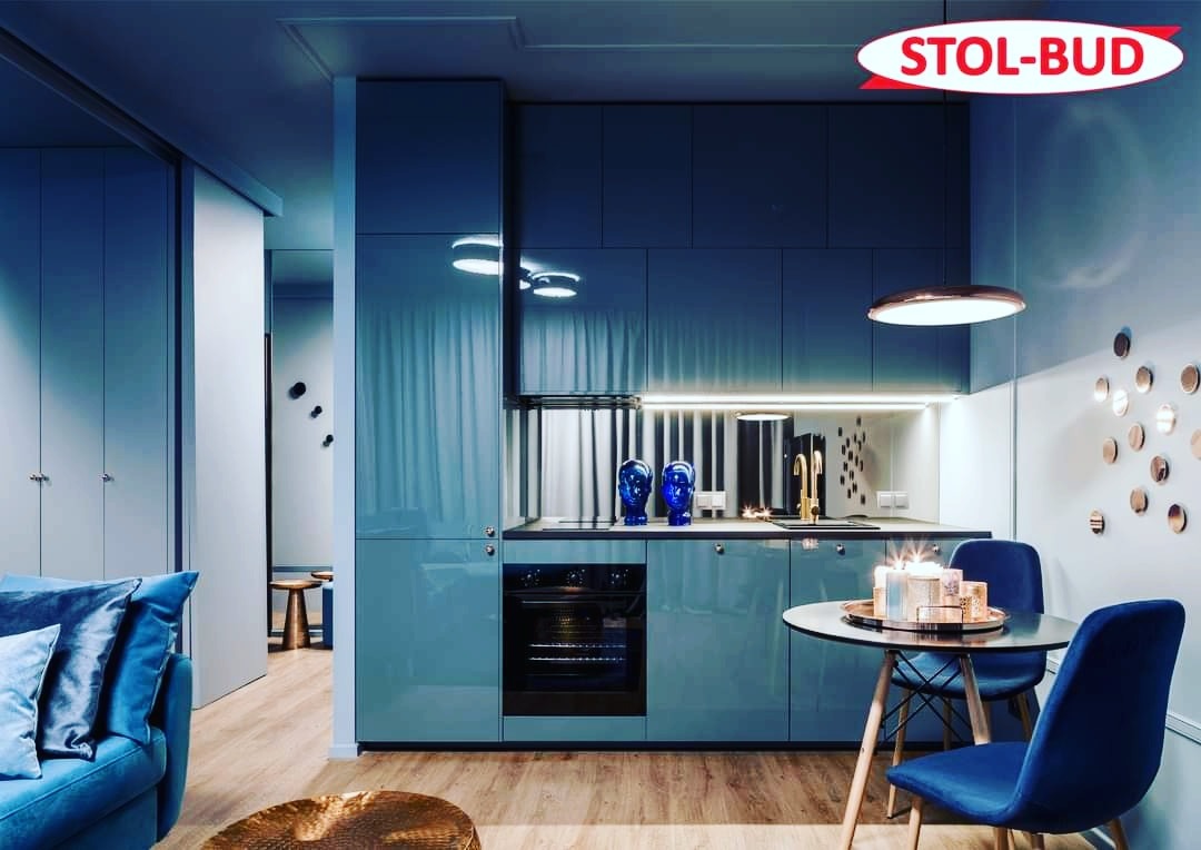 Niebieska, lakierowana zabudowa kuchenna z piekarnikiem, oświetleniem LED i dekoracyjnymi elementami, widoczny stół z krzesłami i logo firmy Stol-Bud w prawym górnym rogu.