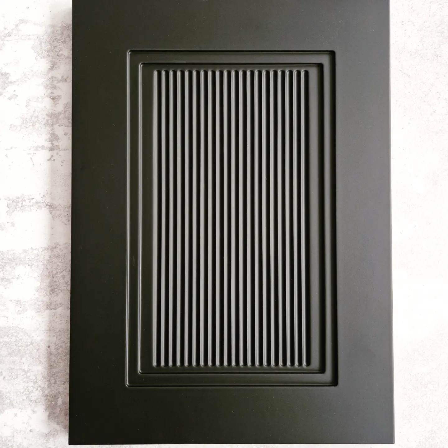 Matowa, czarna frontowa płyta meblowa z frezowanymi, pionowymi żłobieniami w prostokątnej ramce, na tle szarej ściany.