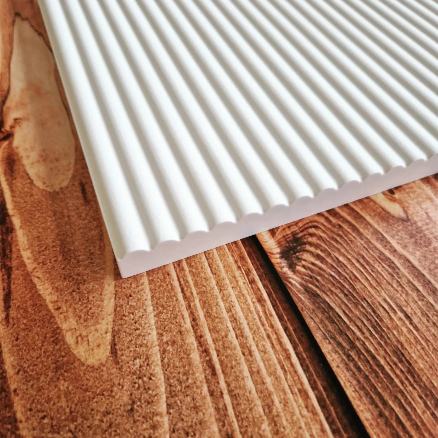 Fragment białego, ryflowanego panelu dekoracyjnego ułożonego na drewnianym blacie o wyraźnym usłojeniu.