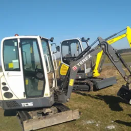 Dwie mini koparki (TC16 i Terex) stoją na trawiastym terenie pod błękitnym niebem z widoczną smugą kondensacyjną samolotu.