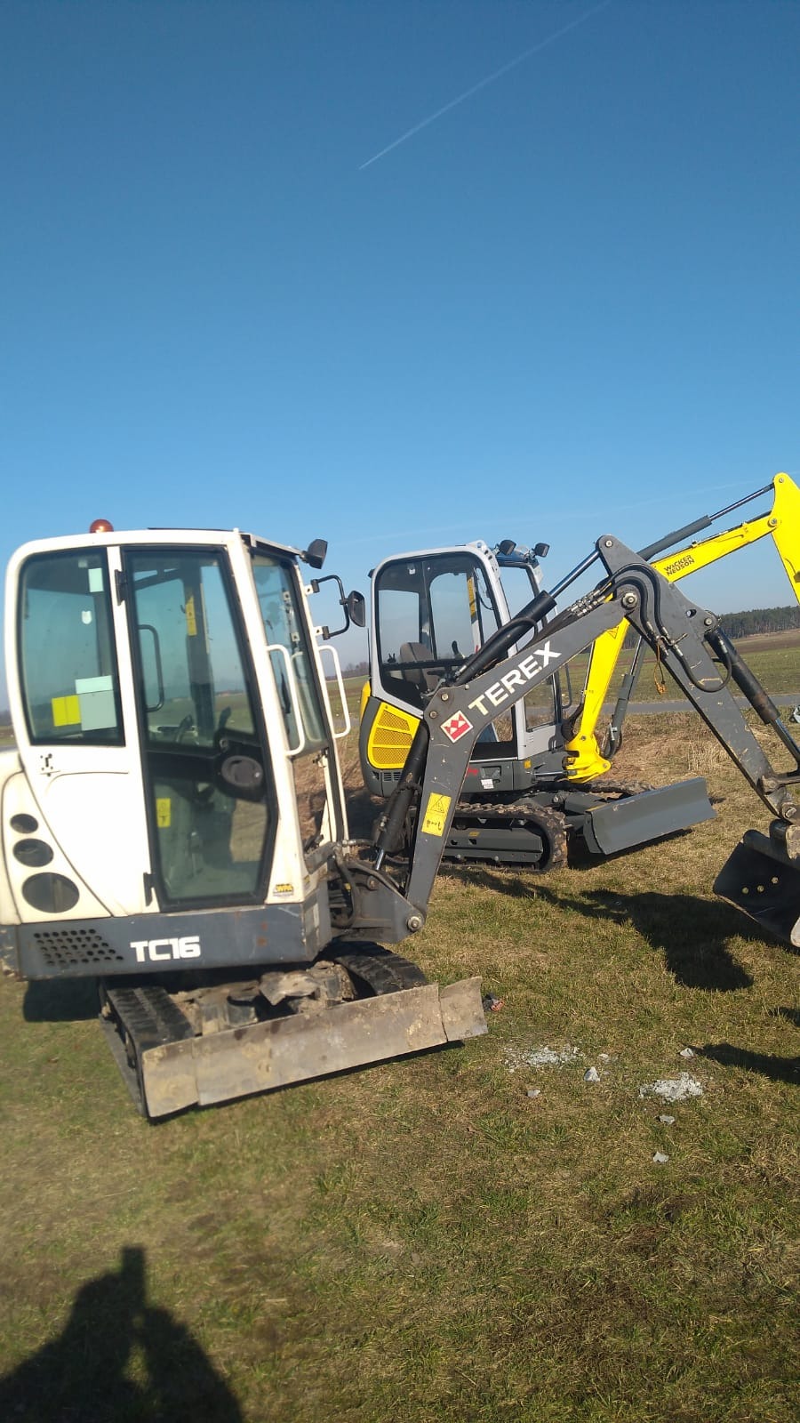 Dwie mini koparki (TC16 i Terex) stoją na trawiastym terenie pod błękitnym niebem z widoczną smugą kondensacyjną samolotu.