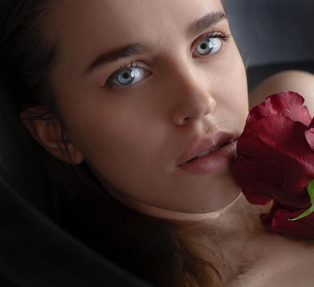 Portret kobiety z intensywnie niebieskimi oczami, kroplami wody na twarzy i czerwoną różą przy ustach, w ciemnym otoczeniu.