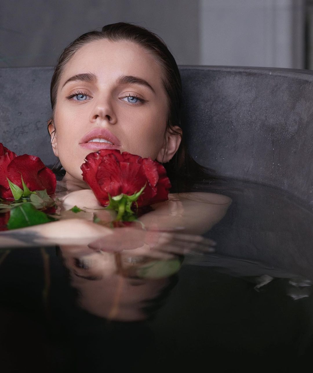 Portret kobiety z niebieskimi oczami w wannie wypełnionej wodą z czerwonymi różami, z widocznym tatuażem na ramieniu.