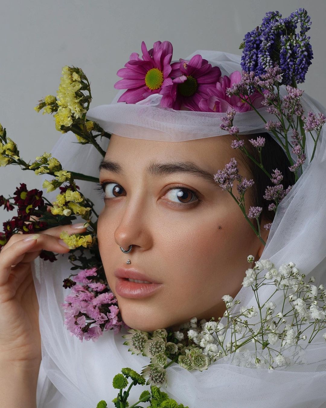 Portret kobiety z piercingiem w nosie i ustach, otoczonej różnorodnymi kwiatami i delikatnym białym woalem.