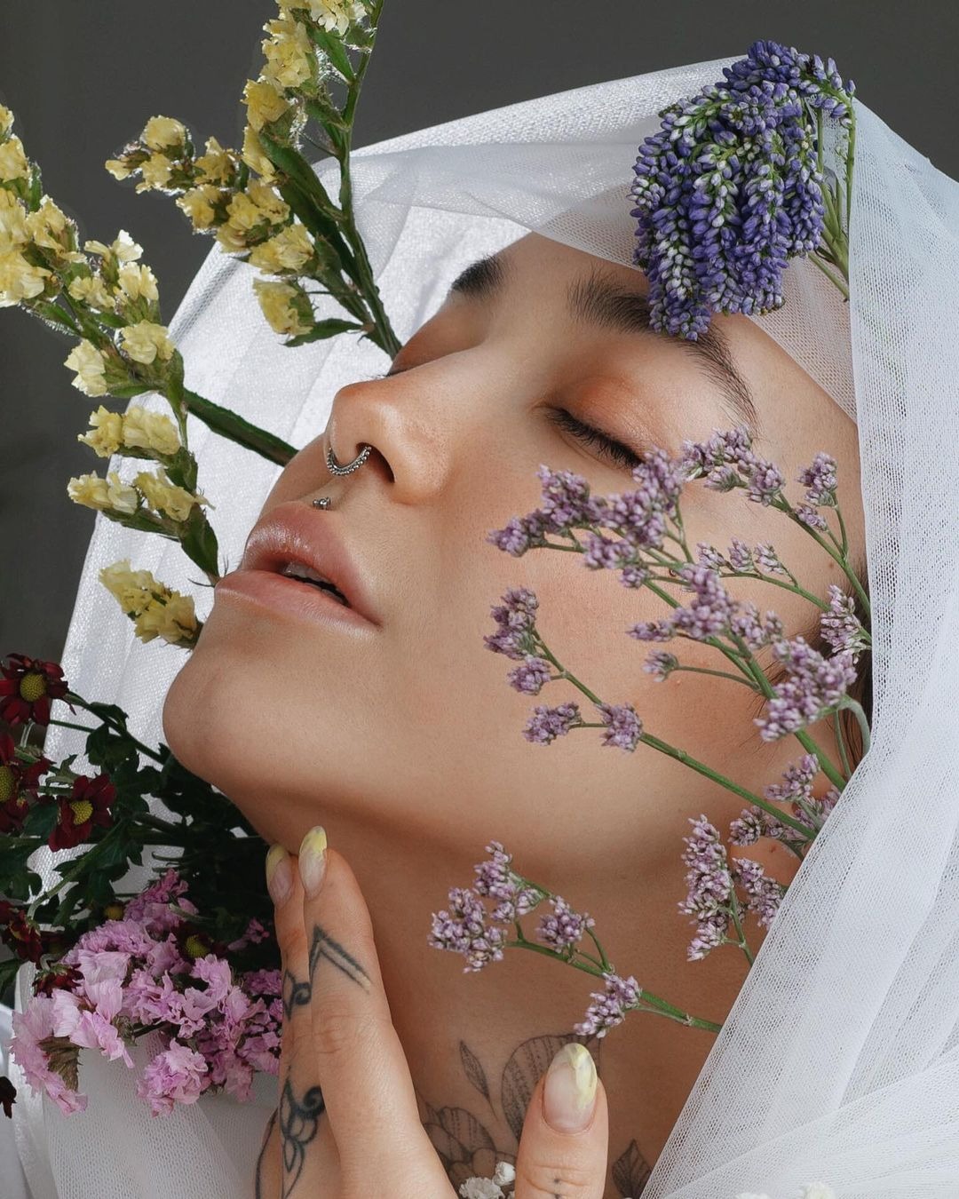 Portret kobiety z zamkniętymi oczami, otoczonej różnokolorowymi kwiatami i delikatną białą tkaniną, z widocznym piercingiem w nosie i tatuażami na dłoni i szyi.