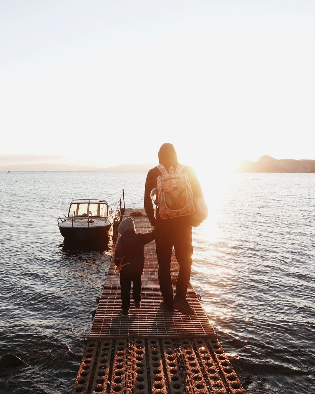 Ojciec z dzieckiem idą pomostem w stronę łodzi na tle zachodzącego słońca nad morzem.