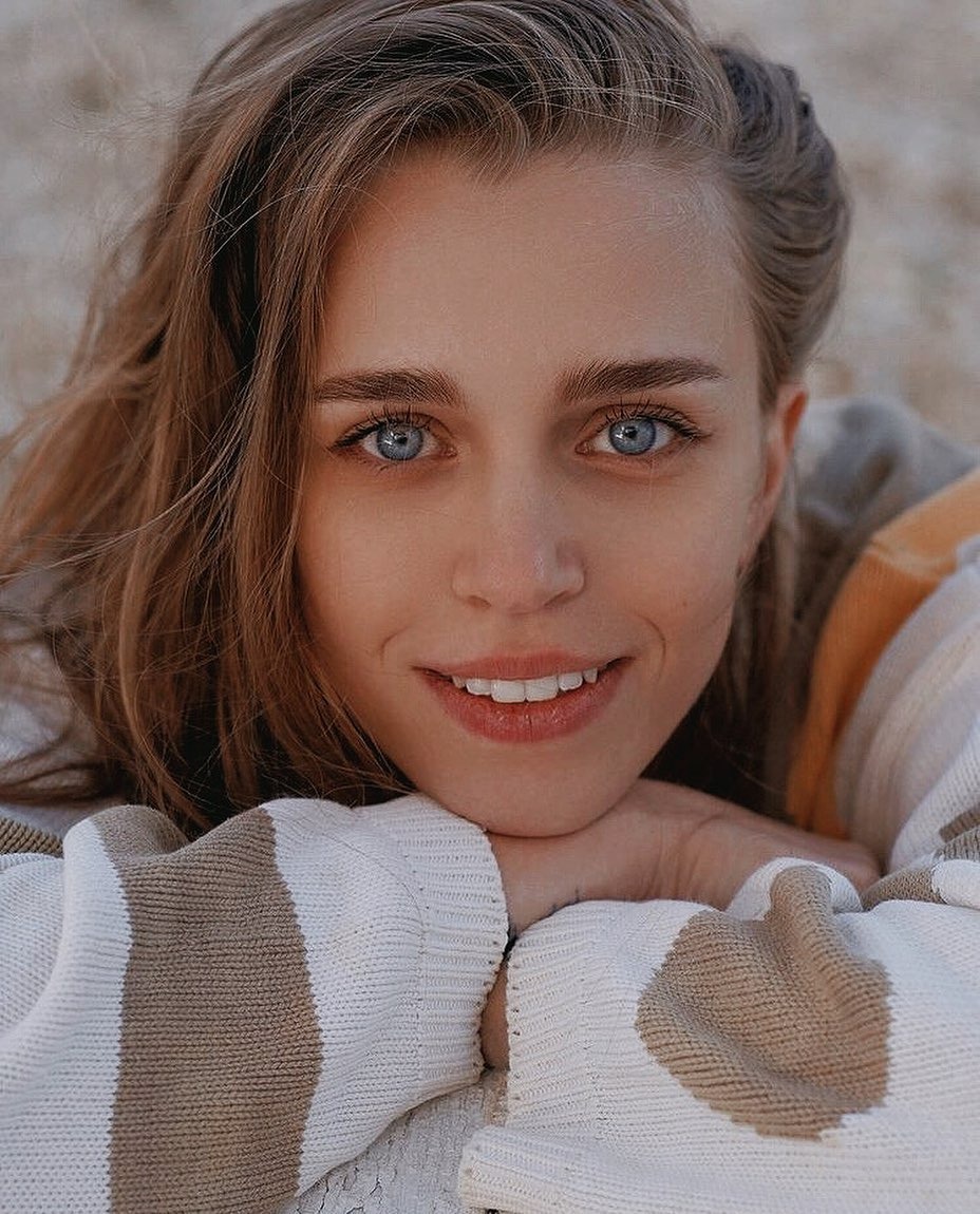 Portret uśmiechniętej młodej kobiety z niebieskimi oczami, opierającej brodę na dłoniach w swetrze w paski.