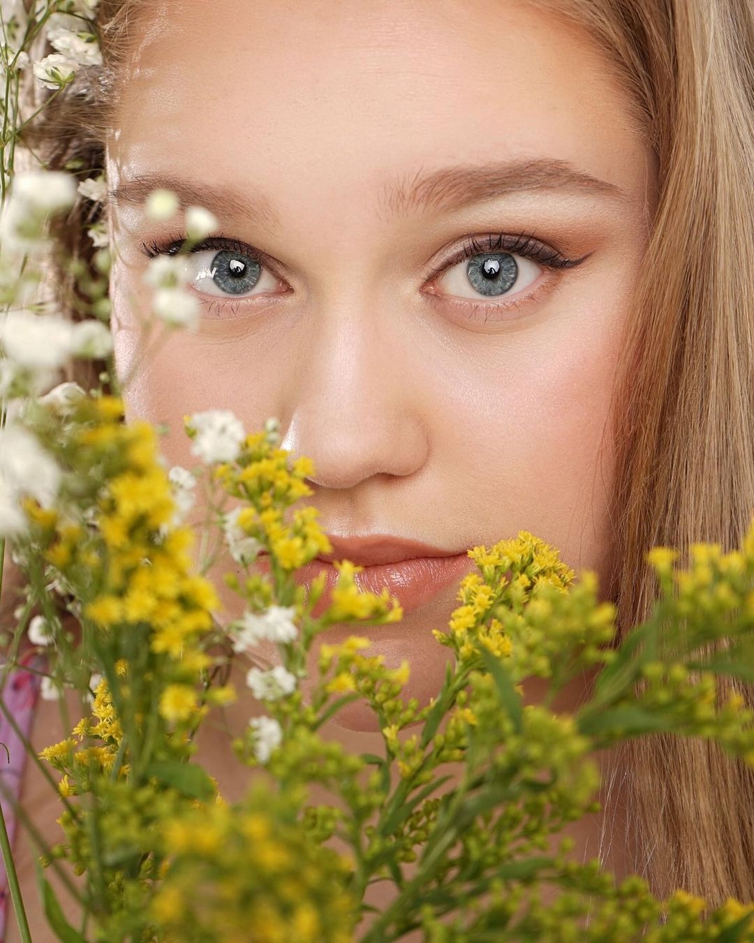 Portret młodej kobiety z niebieskimi oczami, otoczonej bukietem żółto-białych kwiatów, z delikatnym makijażem i blond włosami.