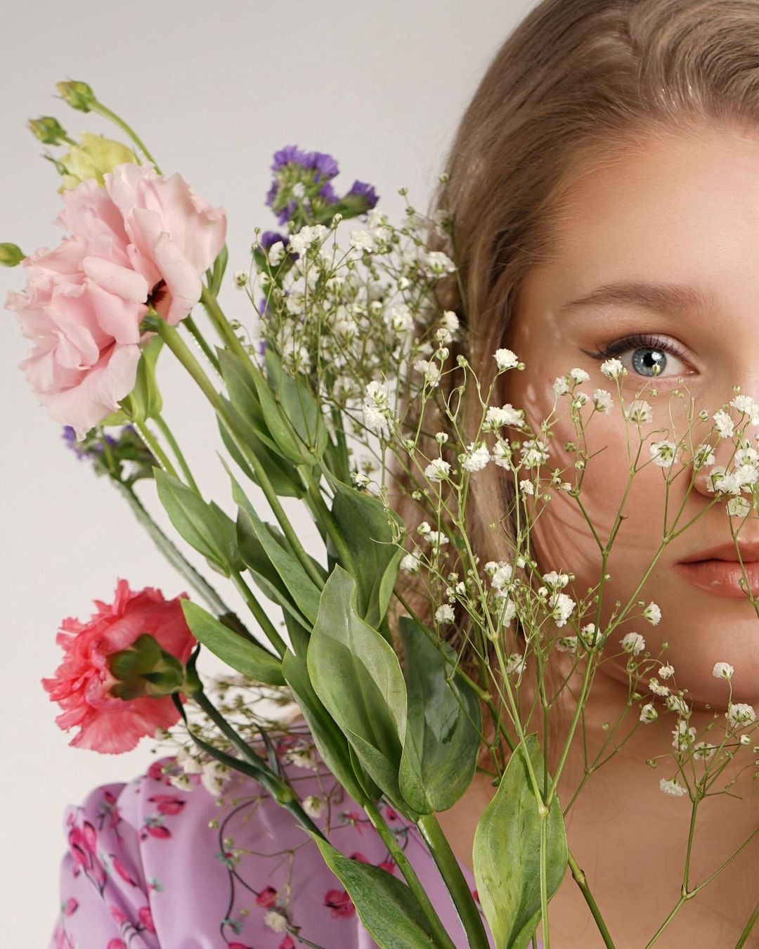 Portret kobiety z bukietem kwiatów, skupiający się na jej niebieskim oku i delikatnym makijażu, z kwiatami częściowo zasłaniającymi twarz.