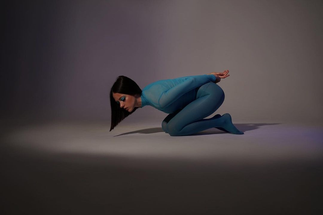 Kobieta w turkusowym stroju body na kolanach w studiu, z rękami splecionymi za plecami i głową skierowaną w dół, oświetlona punktowo.