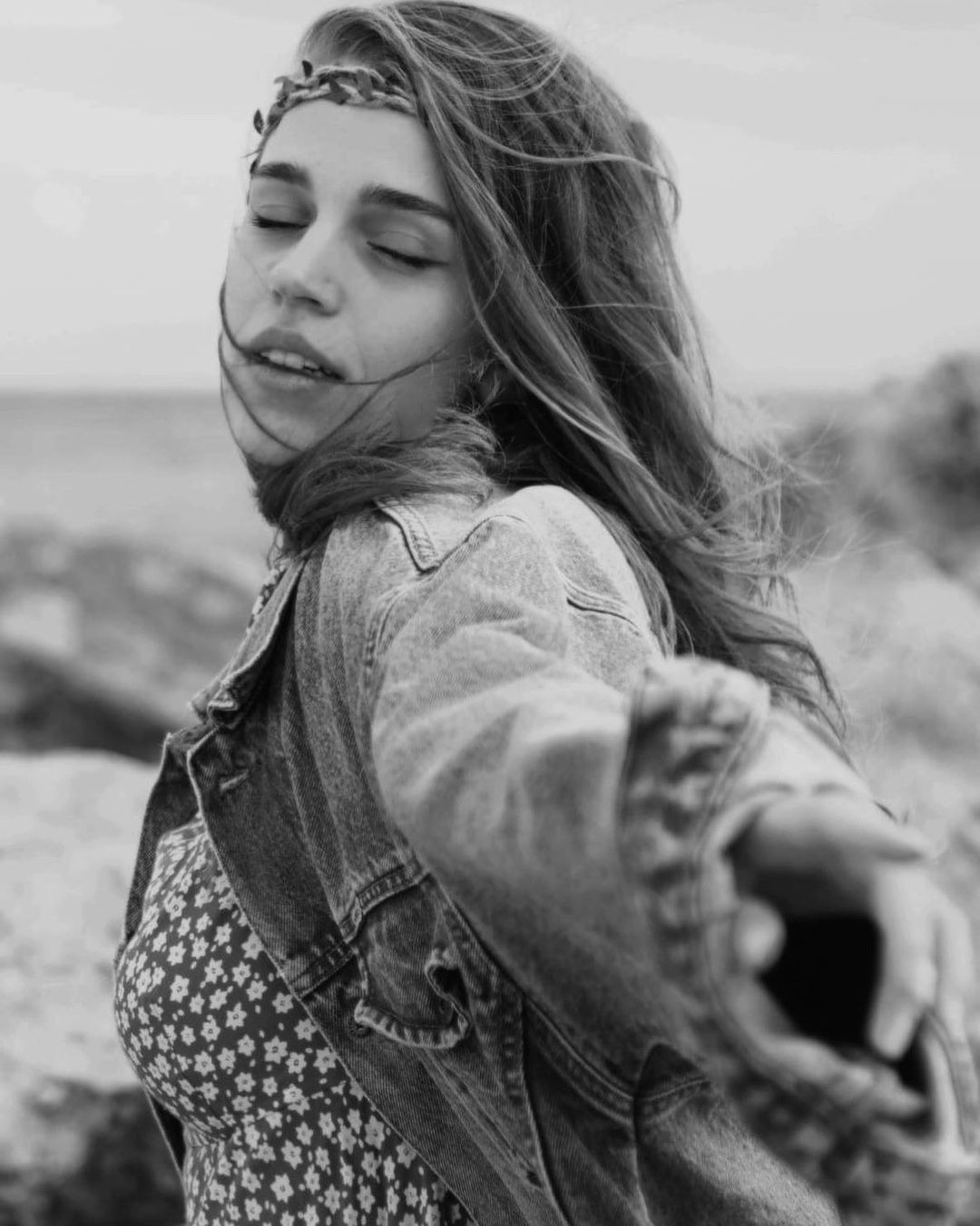 Czarno-biała fotografia portretowa kobiety z zamkniętymi oczami, w dżinsowej kurtce i sukience w kwiaty, z wiatrem we włosach i opaską na głowie, na tle kamienistego wybrzeża.