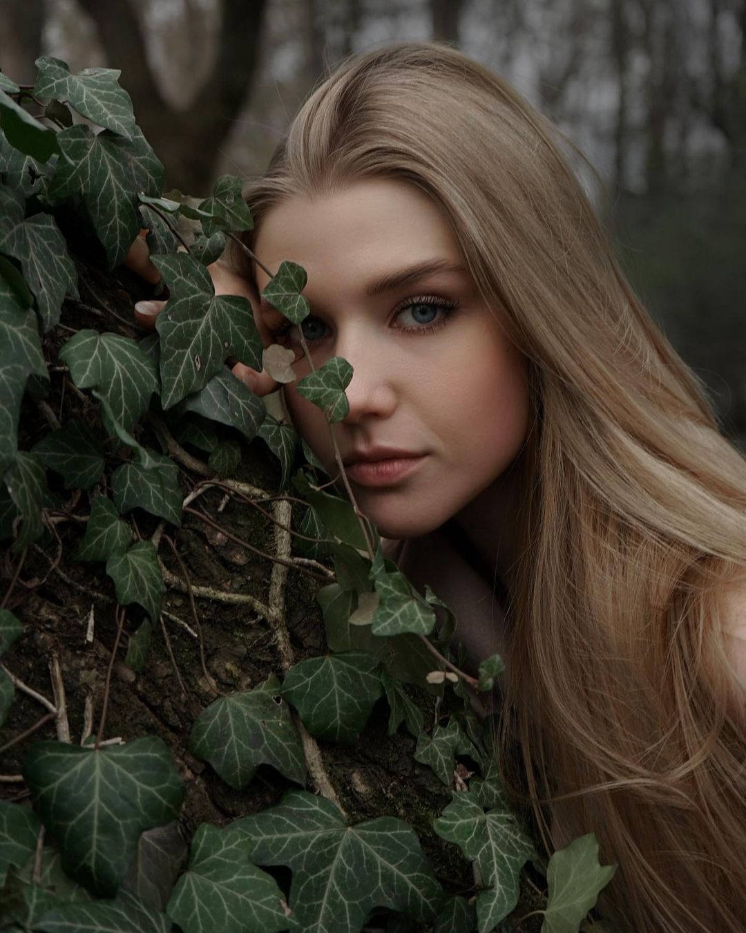 Portret młodej kobiety o blond włosach i niebieskich oczach, opierającej się o pień drzewa porośnięty bluszczem, z delikatnym uśmiechem.