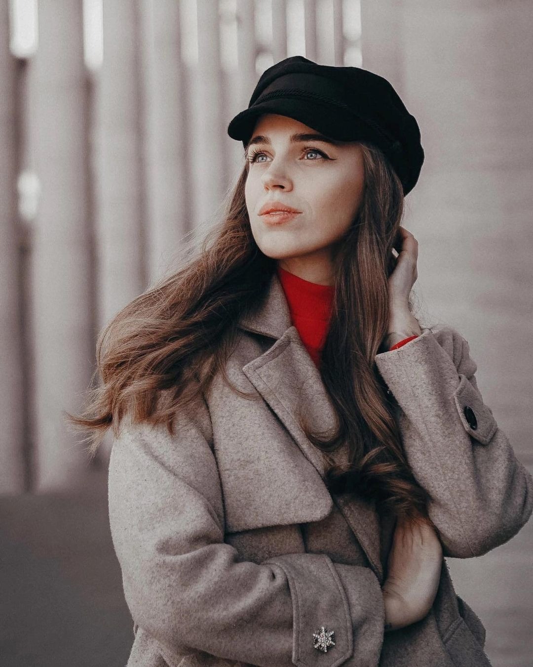 Portret kobiety w płaszczu i czapce, z ręką we włosach, spoglądającej w lewo, na tle jasnych pionowych linii.