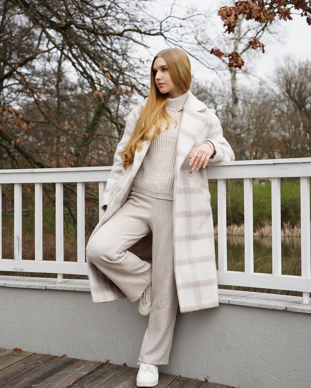 Blondwłosa kobieta w beżowym płaszczu w kratę i spodniach, oparta o białą barierkę z widokiem na staw w tle.