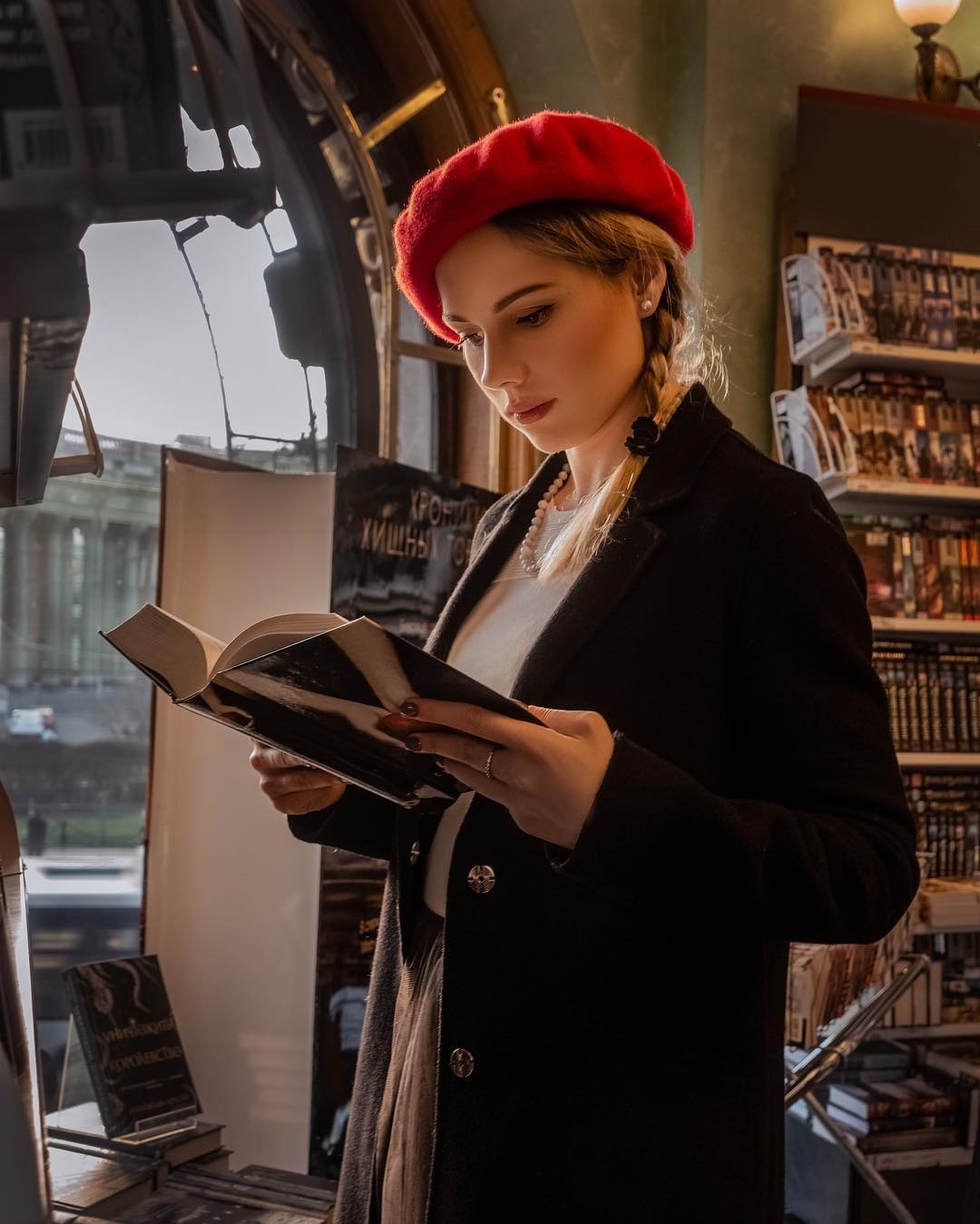 Kobieta w czerwonym berecie czyta książkę w księgarni, przy oknie z widokiem na ulicę.