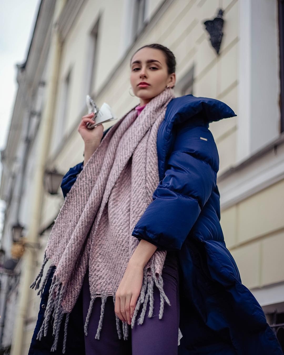 Portret kobiety w niebieskiej kurtce i różowym szalu, trzymającej w dłoni złożoną gazetę, z budynkiem w tle, sfotografowany z dołu.