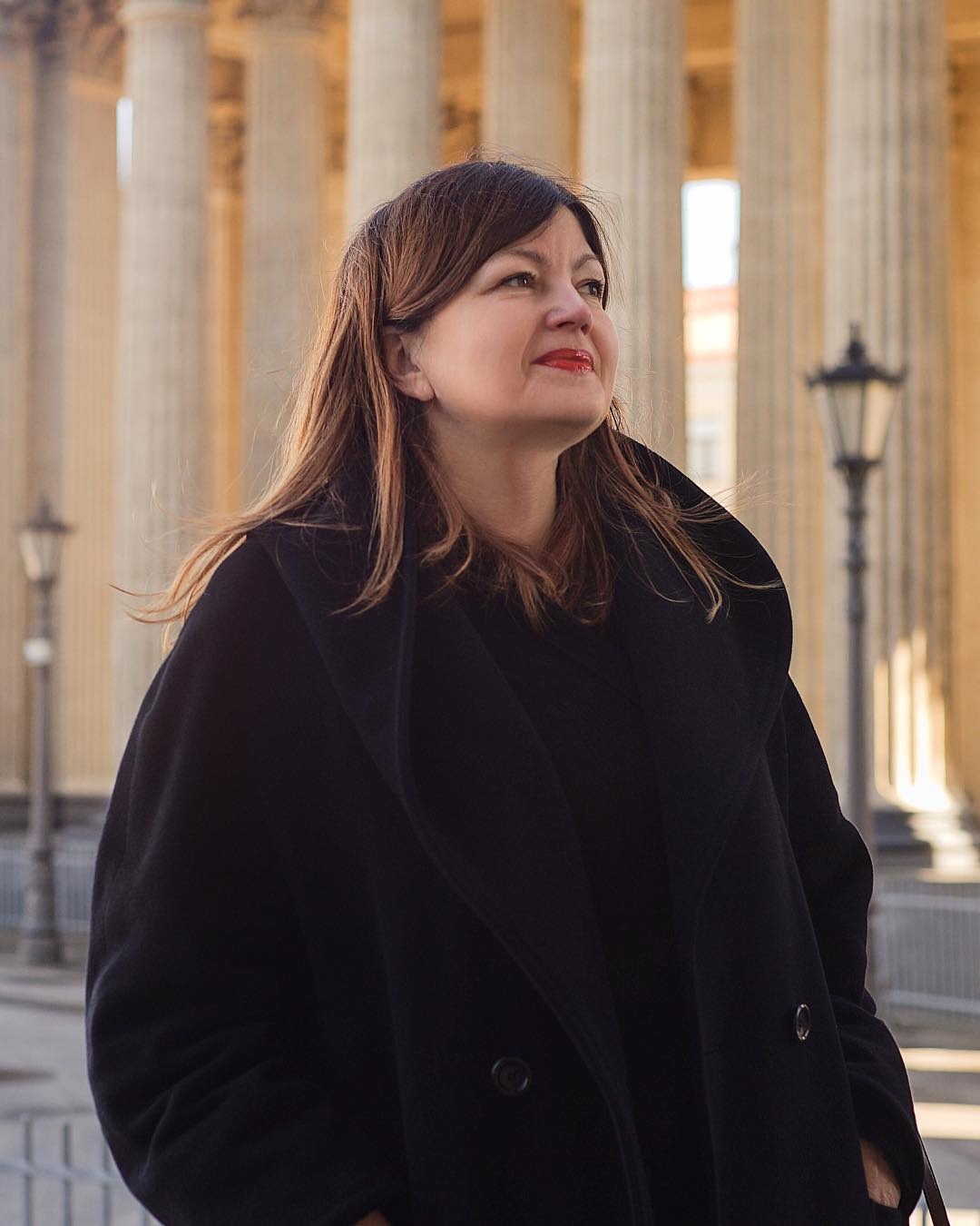 Portret kobiety w czarnym płaszczu na tle kolumn i latarni, z lekkim uśmiechem i spojrzeniem w górę.