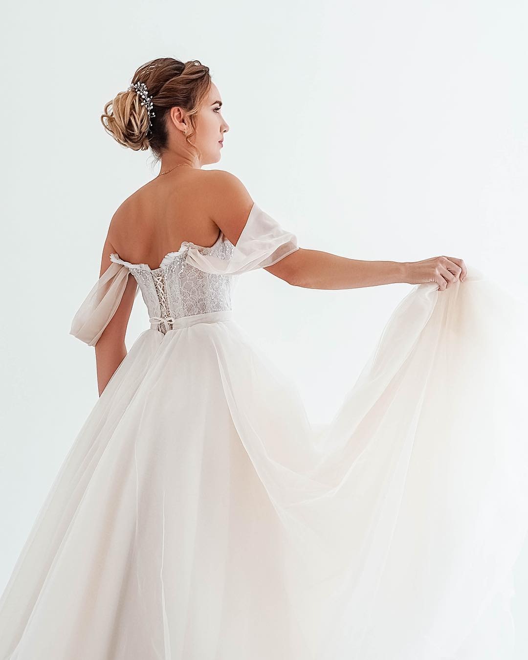 Panna młoda w białej sukni z odkrytymi ramionami i delikatnym koronkowym gorsetem, upięcie włosów z ozdobną spinką, trzyma falujący dół sukni, widok z tyłu, na jasnym tle.