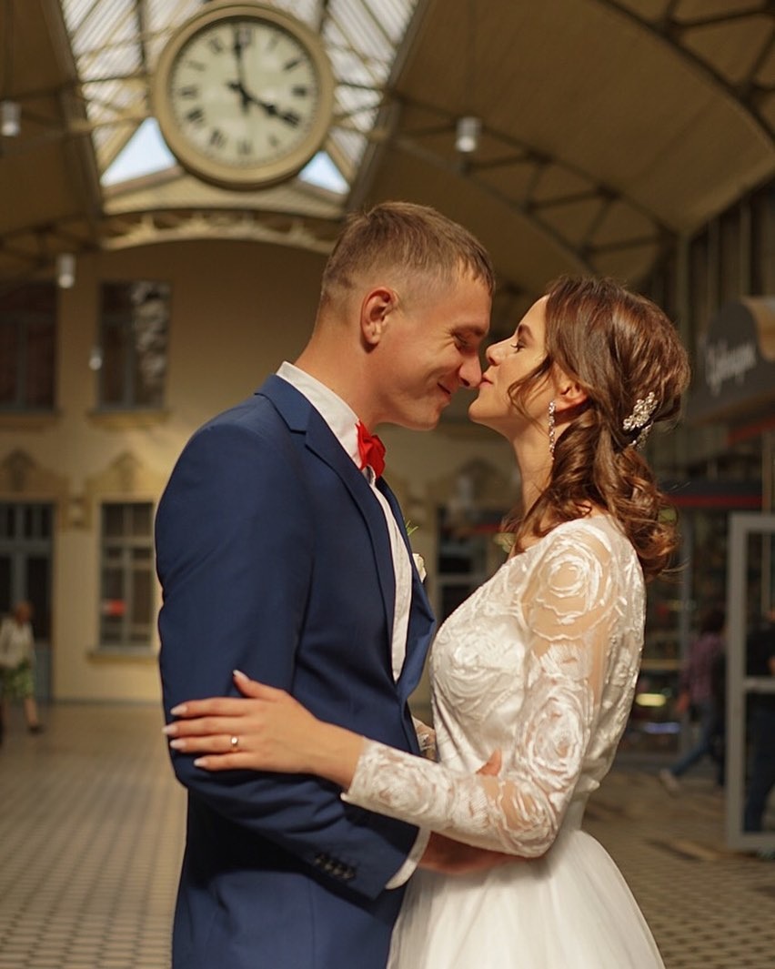 Para narzeczonych w eleganckich strojach, on w granatowym garniturze z czerwoną muchą, ona w białej sukni z koronkowymi rękawami, tulą się do siebie na tle hali dworcowej z dużym zegarem w Poznaniu.