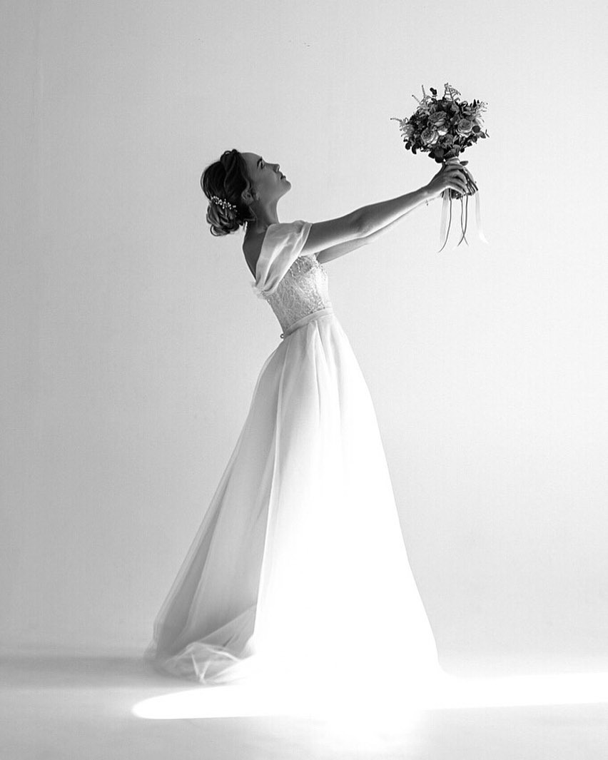 Czarno-białe zdjęcie kobiety w sukni ślubnej, unoszącej bukiet kwiatów na wyciągniętych rękach, z głową skierowaną ku górze, na jasnym tle.