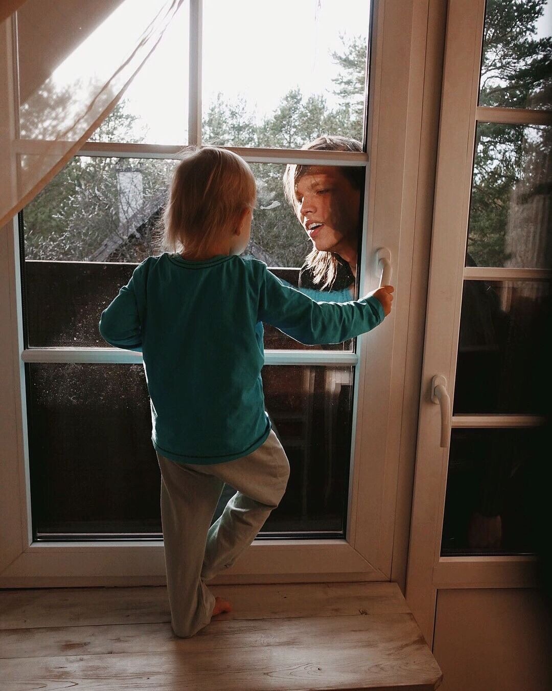 Dziecko w turkusowej koszulce wspina się na parapet, trzymając się klamki okna, podczas gdy twarz dorosłego jest widoczna przez szybę z zewnątrz.