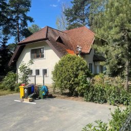 Mycie ciśnieniowe - Pracownik w białym kombinezonie czyści dach domu z czerwonej dachówki za pomocą myjki ciśnieniowej. Widoczne zabrudzenia na dachu i otoczenie domu.