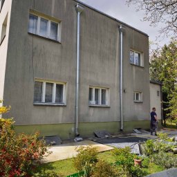 Mycie ciśnieniowe - Szara elewacja dwukondygnacyjnego budynku z rynnami, widoczna z poziomu ogrodu. Mężczyzna w tle, prawdopodobnie wykonawca, sugeruje prace konserwacyjne lub czyszczenie.