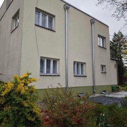 Mycie ciśnieniowe - Dwupiętrowy budynek z beżową elewacją i srebrnymi rynnami, widoczny z poziomu ogrodu z kwitnącymi krzewami. Ujęcie z boku, w pochmurny dzień.