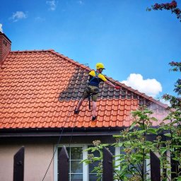 Mycie ciśnieniowe - Pracownik w kasku i uprzęży czyści czerwoną dachówkę z zabrudzeń za pomocą lancy ciśnieniowej. Widoczny fragment dachu przed i po czyszczeniu. W tle błękitne niebo.