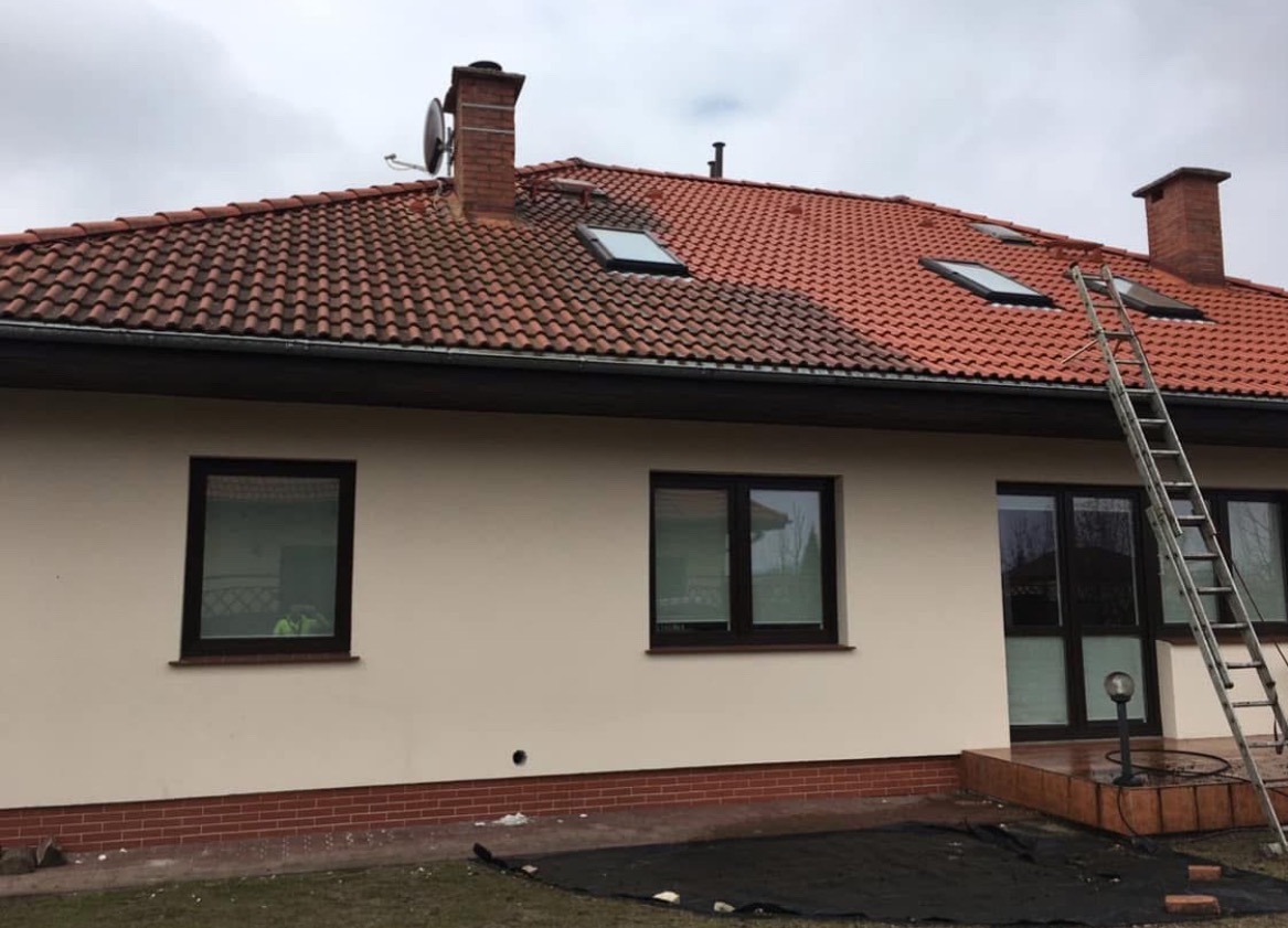 Częściowo umyty dach z czerwonej dachówki, widoczny kontrast czystej i zabrudzonej powierzchni, drabina oparta o ścianę domu z kremową elewacją i brązowymi oknami, fragment czarnej plandeki...