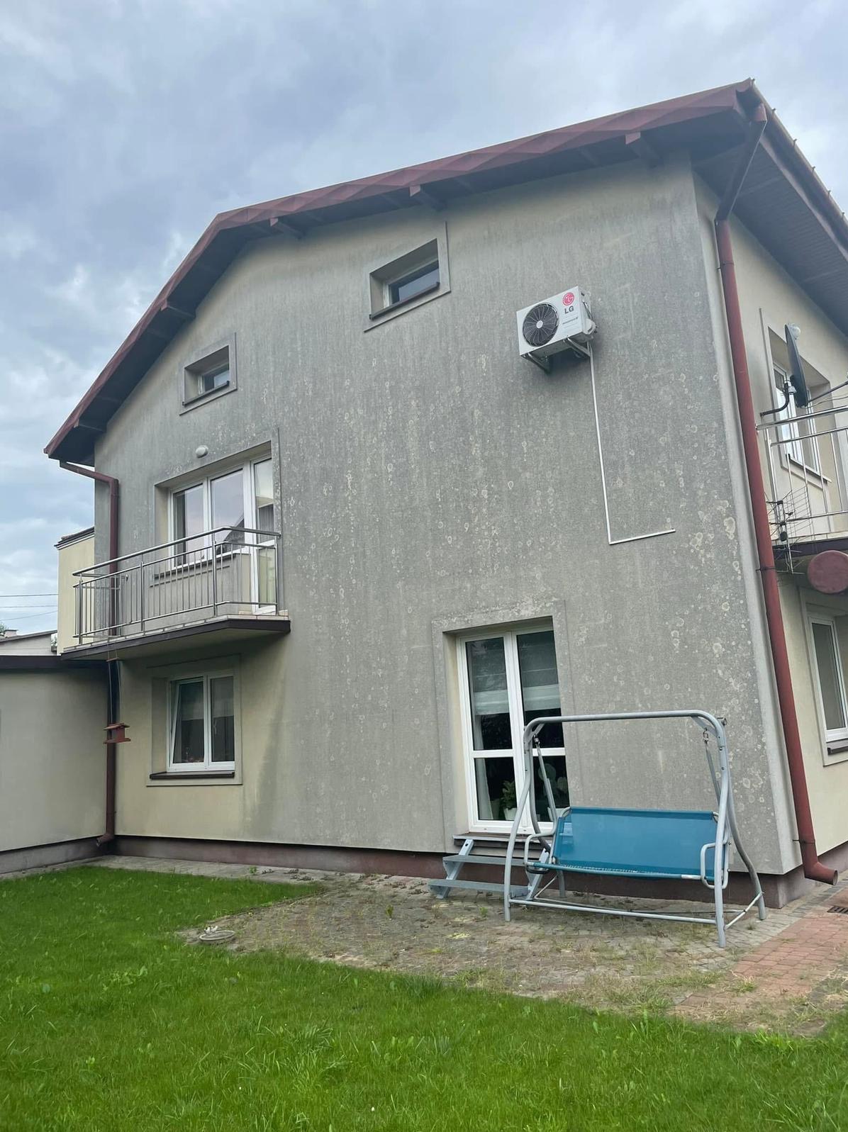 Dwupiętrowy dom z szarą elewacją, balkonem i klimatyzatorem LG. Widoczne zabrudzenia na ścianie. Na pierwszym planie huśtawka ogrodowa i trawnik.